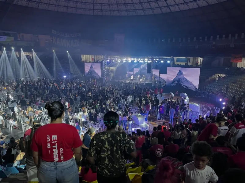 Público em pé diante de um palco iluminado em ginásio, com telões nas laterais
